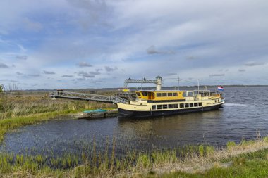 Lauwersmeer Gölü 'ndeki rıhtımdan kalkan sarı yolcu feribotu bulutlu gökyüzü altında, Friesland, Hollanda