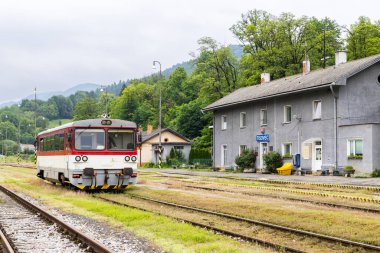 Slovakya 'nın Tisovec istasyonunda yeşil doğayla çevrili kırmızı ve beyaz tren bekliyor.