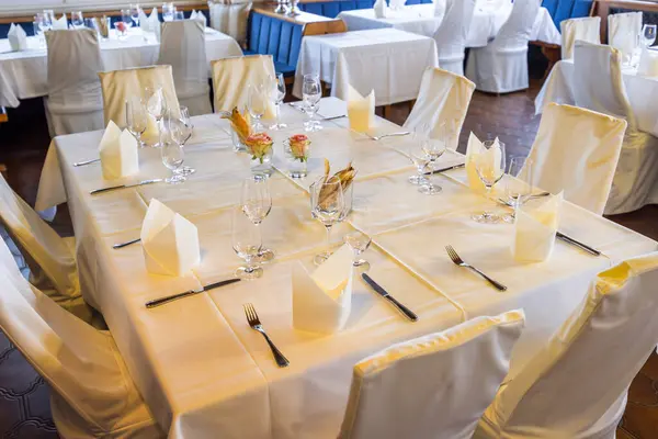 Lüks restoranda beyaz masa örtülü, peçeteli, çatal bıçaklı, bardaklı ve çiçekli konukları bekleyen zarif bir masa.