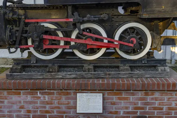 Eski bir buharlı lokomotifin kırmızı bağlantı çubukları ve beyaz tekerlekleri tuğla kaidede sergileniyordu.