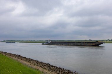 Hollanda, Alblasserdam 'da bulutlu bir gökyüzünün altında geniş bir nehir boyunca büyük bir mavnayı iten römorkör.