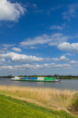 Hollanda, Utrecht 'teki Lopik yakınlarındaki Lek Nehri' nde konteynır taşıyan iç kargo gemisi, beyaz bulutlu mavi bir gökyüzünün altında,