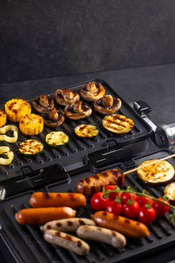Lezzetli ızgara sosisler, mantarlar, kabak, mısır ve domatesler elektrikli ızgarada pişiyor.
