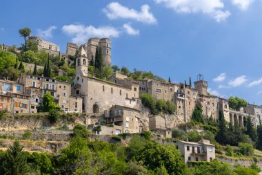 Montbrun les Bains 'in, ortaçağ mimarisi ve kalesinin kalıntılarını güneşli bir gökyüzünün altında sergilediği bir tepenin manzarası.