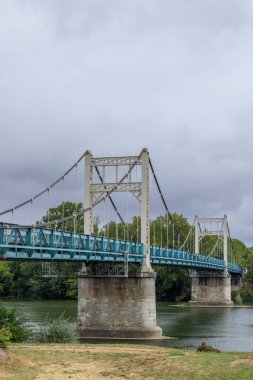 Auvillar, Occitanie, Fransa 'da, bulutlu bir gökyüzünün altında Garonne nehrinden geçen turkuaz güverteli metal asma köprüsü.