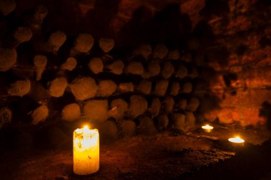 Karanlık bir mahzende yakacak odun yığınıyla yanan beyaz mum sıcak ve atmosferik bir manzara yaratıyor.