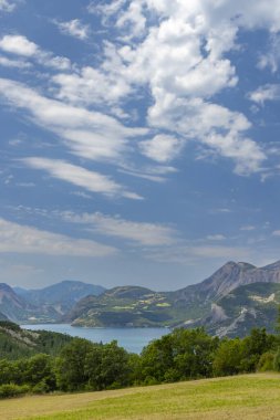 Serre Poncon Gölü 'nün nefes kesen manzarası Hautes Alpes' te, Fransız Alplerinde bulutlarla dolu canlı mavi bir gökyüzünün altındaki dağların arasına yerleşmiş.