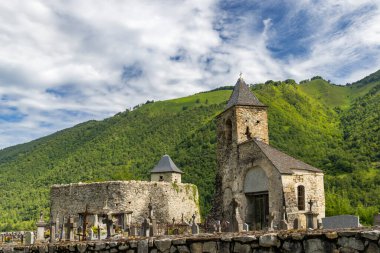 Ustou, Ariege, Occitanie, Fransa 'daki yemyeşil dağların zemininde eski bir kilise ve mezarlığın yıpranmış mezar taşları ve haçları görülüyor.