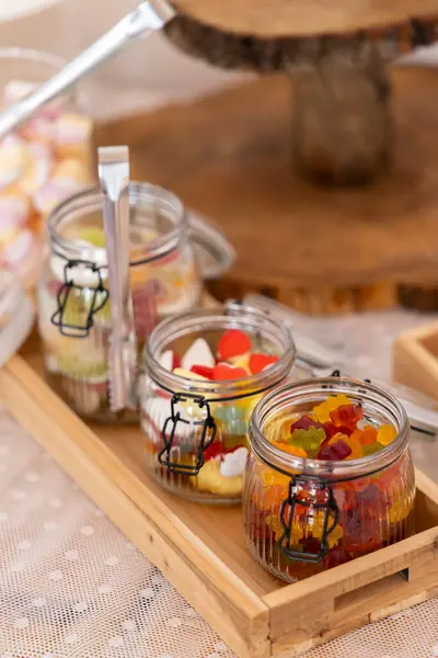 Cam kavanozlar çeşitli renkli şeker ve maşalarla dolu, ahşap tepside dinleniyor, tatlı bir ikram için hazır.