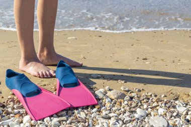 Çakıl taşı plajında, pembe ve mavi paletlerin yanında duran çıplak ayaklı çocuk, şnorkelle yüzmeye hazırlanıyor.