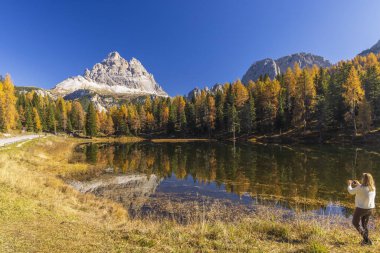 Tre Cime di Lavaredo 'nun fotoğraflarını çeken turist, İtalya' da Auronzo di Cadore, Dolomites 'de sonbahar yeşillikleri sırasında Antorno Gölü' ne yansıyor.