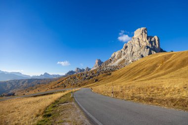 Güneşli bir sonbahar gününde İtalyan Dolomitleri 'nde Passo Giau' nun heybetli dolomit tepelerine doğru altın çayırlar boyunca uzanan manzaralı dolambaçlı yol.