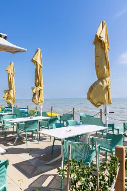 Güneşli bir yaz gününde Macaristan 'ın Balaton Gölü yakınlarındaki bir açık hava restoranında kapalı sarı şemsiyeler ve turkuaz sandalyeler ve beyaz masalar.