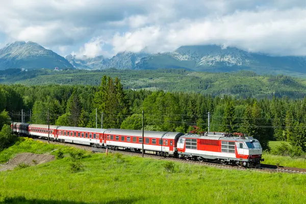 Kırmızı ve beyaz elektrikli yolcu treni, arka plandaki yüksek Tatras dağ sırası olan Slovakya 'daki Strba yeşil vadisinden geçiyor.