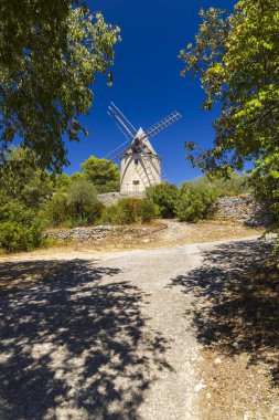 Açık mavi gökyüzünün altındaki ağaçların arasında duran manzaralı yel değirmeni.
