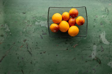 Limonlar ve portakallar desenli yeşil bir yüzey üzerinde siyah bir tel sepetin içinde dizilmişler.