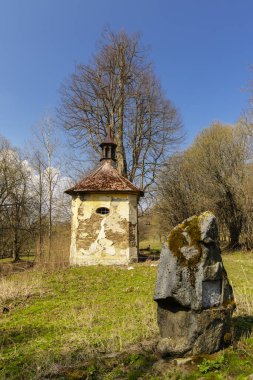 Önünde yosunlu kayalar, baharda bir tarlada, harap olmuş cephesi ve paslı çatısı olan eski bir şapel.
