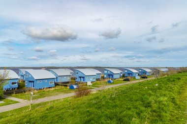 Bulutlu bir gökyüzünün altında gölün kıyısında birkaç mavi yazlık ev duruyor.