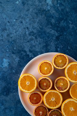 Dilimlenmiş portakallar ve kan portakalları pembe bir tabağa dizilerek dokulu mavi bir zemin üzerinde canlı bir kontrast oluşturuyor.