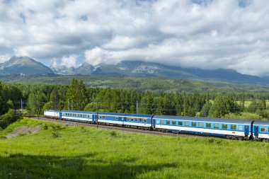 Strba, Slovakya yakınlarındaki yüksek Tatras dağlarıyla yeşil tarlalardan geçen mavi tren.