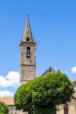 Üzerinde haç olan taş çan kulesi yeşil ağaçlar ve mavi gökyüzünün altında küçük bir Avrupa kasabasında eski binalar.