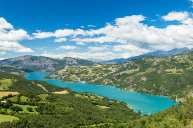 Serre Poncon Gölü 'nün nefes kesen manzarası Fransız Alpleri' nin çerçevelediği yemyeşil vadide süzülüyor ve canlı mavi bulutlarla dolu bir gökyüzünün altında evlerle süsleniyor.