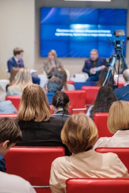 Modern salonda sandalyelerde oturan, hoparlörleri dinleyen ve iş konferansı sırasında sunum izleyen isimsiz insanlar.