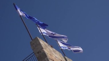 Yazın bulutsuz mavi gökyüzüne doğru rüzgarda dalgalanan İsrail bayraklarıyla yaşlanmış taş duvarın statik çekimlerinden.