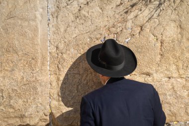 Siyah takım elbiseli ve şapkalı bir adam güneşli bir günde dua ederken kutsal ağlama duvarına bakıyor.
