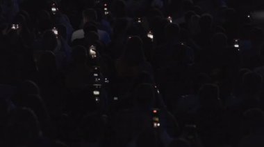 Bir grup izleyicinin cep telefonlarından canlı müzik dinletisi kaydederken yüksek açılı arka plan görüntüsü yanıp sönen ışıkta