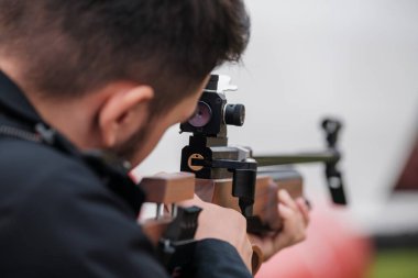 Siyah tişörtlü, tanınmayan erkek profesyonel nişancının arka plan görüşü açık havada bulanık arka planda hedefe şık bir silah doğrultuyor.