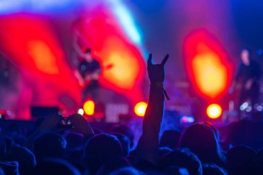Arkaya bakan heyecanlı erkek izleyiciler rock 'n roll işareti gösterirken bulanık gitarist sahnede canlı performans sergiliyor. Aydınlatılmış turuncu ışıklarla seyircileri alkışlıyor.