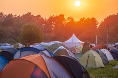 Kırsal kesimdeki müzik festivali sırasında yeşil ağaçların yanına gün batımına karşı kamp çadırları kurulmuş.