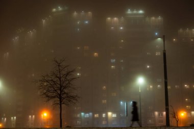 Yoğun sisli bir gecede apartman binası ve yürüyen insan silüetiyle parlayan fenerler.
