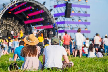 Güneşli bir yaz günü müzik festivali sırasında kalabalık ve sahnenin önünde sıradan giysiler içinde tanınmayan bir çiftin arka planda oturuşu.
