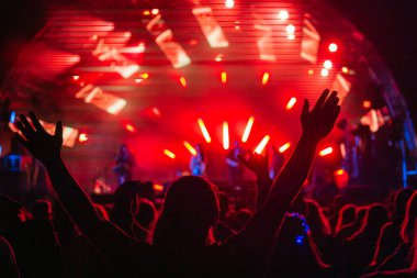 Arka plan silueti, kolları havada müzik festivalinin ve sahnedeki parlak kırmızı ışıkla bando performansının keyfini çıkaran insanların görüntüsü.