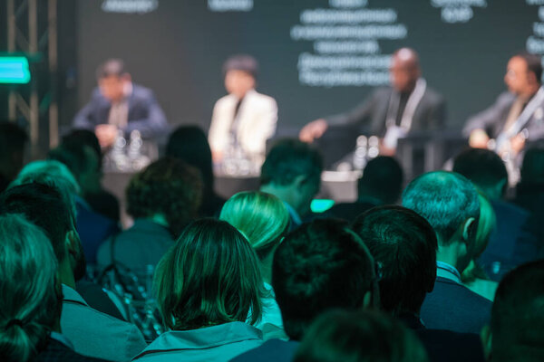 Толпа профессионалов, присутствующих на бизнес-конференции, с акцентом на докладчиков в панельной дискуссии. Заинтересованная аудитория внимательно слушает лидеров отрасли, которые делятся своими идеями.