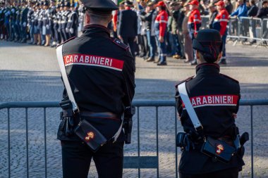 Two Italian carabinieri officers in uniform observe a public event. They stand alert behind a barricade, focusing on crowd security during a formal gathering. clipart