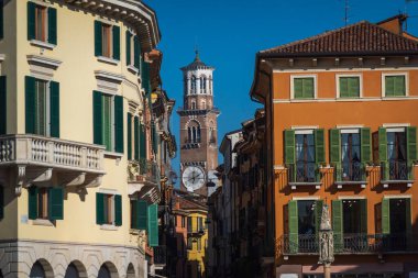 Verona 'daki sokak manzarası, canlı binalar ve mavi gökyüzünün altında antik saat kulesi. İtalya 'nın mimari cazibesini ve tarihini yakalıyor.