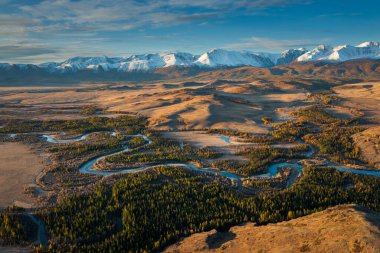 Altından sonbahar çayırlarında akan nehrin havadan görünüşü ormanlık vadi, arka planda karlı dağ sıraları, canlı huzurlu doğal manzara açık hava keşfi ve doğa takdiri için mükemmel.