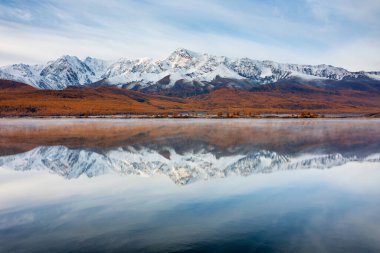 Sakin göle yansıyan karla kaplı nefes kesici dağ sıraları altın sonbahar ormanı ve parlak mavi gökyüzü ile çevrili.