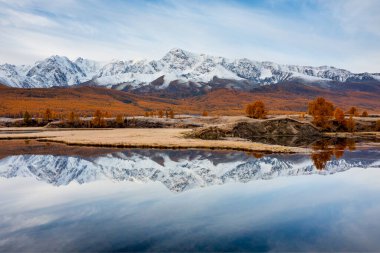 Huzurlu göl yansıması ve önplanında sonbahar ağaçları olan güzel karlı dağ sıraları. Sonbahar dağ güzelliği ve sükuneti yakalamak için mükemmel bir doğal manzara..