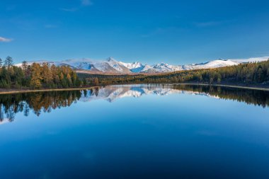 Dağ gölü. Mavi gökyüzünün altındaki karlı tepelerin ve sonbahar ağaçlarının kristal berraklığında yansıması. Huzurlu ve doğal manzara meditasyon ve uzak manzaralar için ideal.. 