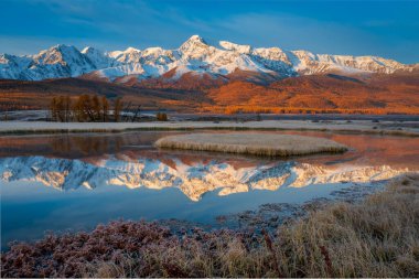 Majestic autumn landscape featuring snow-capped mountains under blue sky reflected on calm water of an alpine lake with golden meadows. clipart