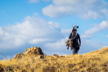 Çimenli ovalarda kamera ekipmanları taşıyan bir fotoğrafçının yürüdüğü açık hava sahnesi. Parlak berrak gökyüzü ve doğal çevre, evcilleştirilmemiş ortamda özgürlük keşfi ve yaratıcılığı çağrıştırıyor.