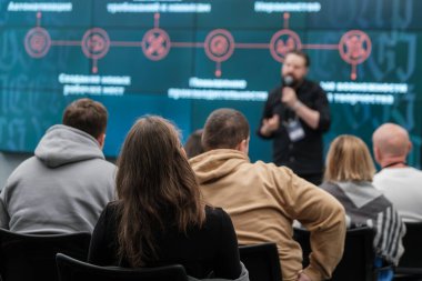 Konferans ortamında konuşmacı dinleyen bir grup insan. Seyirciler sunum slaytlarına odaklandı.