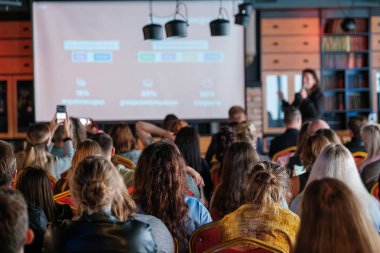 Seyirciler konferans odasında oturmuş ekranda görüntülerini sunan bir konuşmacıyı dinliyorlar..