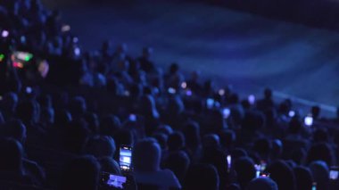 Kalabalık bir tiyatro seyircisi, loş ışıklandırmalı akıllı telefonlu canlı bir sahne etkinliğini filme alıyor..