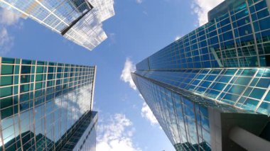 Uzun, modern cam gökdelenler mavi gökyüzüne uzanıyor, bulutları yansıtıyor, dinamik perspektif yaratıyor.