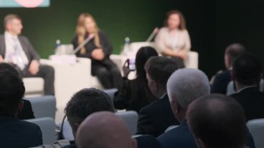 Bir panel tartışması sırasında konuşmacıları dinlerken konferansa katılan iş adamları. Şirket temalarını vurgulayan bir arkaplan ve ağ oluşturma içerir.